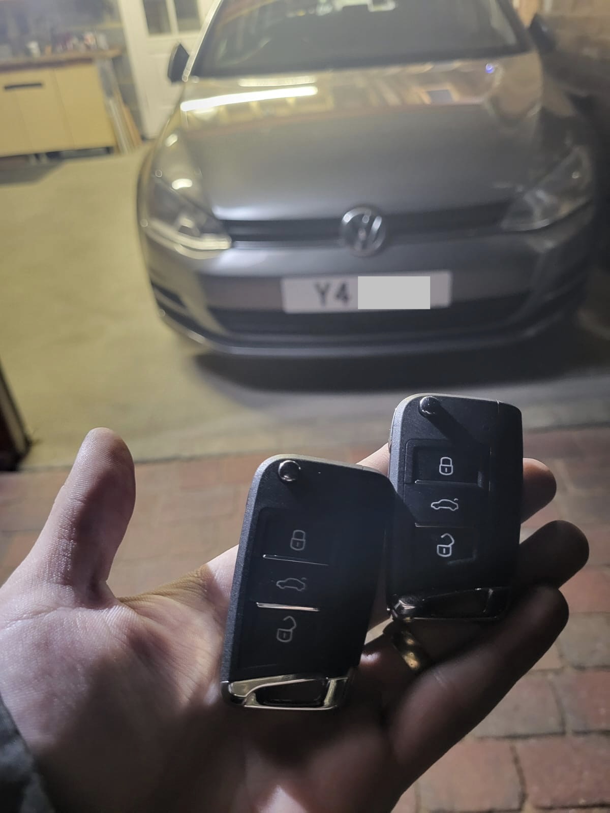 Two Volkswagen flip remote keys held in a hand in front of a grey Volkswagen Golf parked in a garage