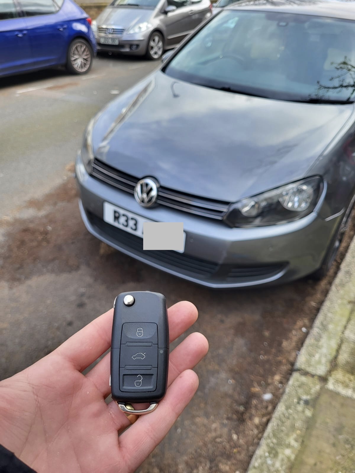 Volkswagen three-button flip key held in front of a grey VW Golf in a car park
