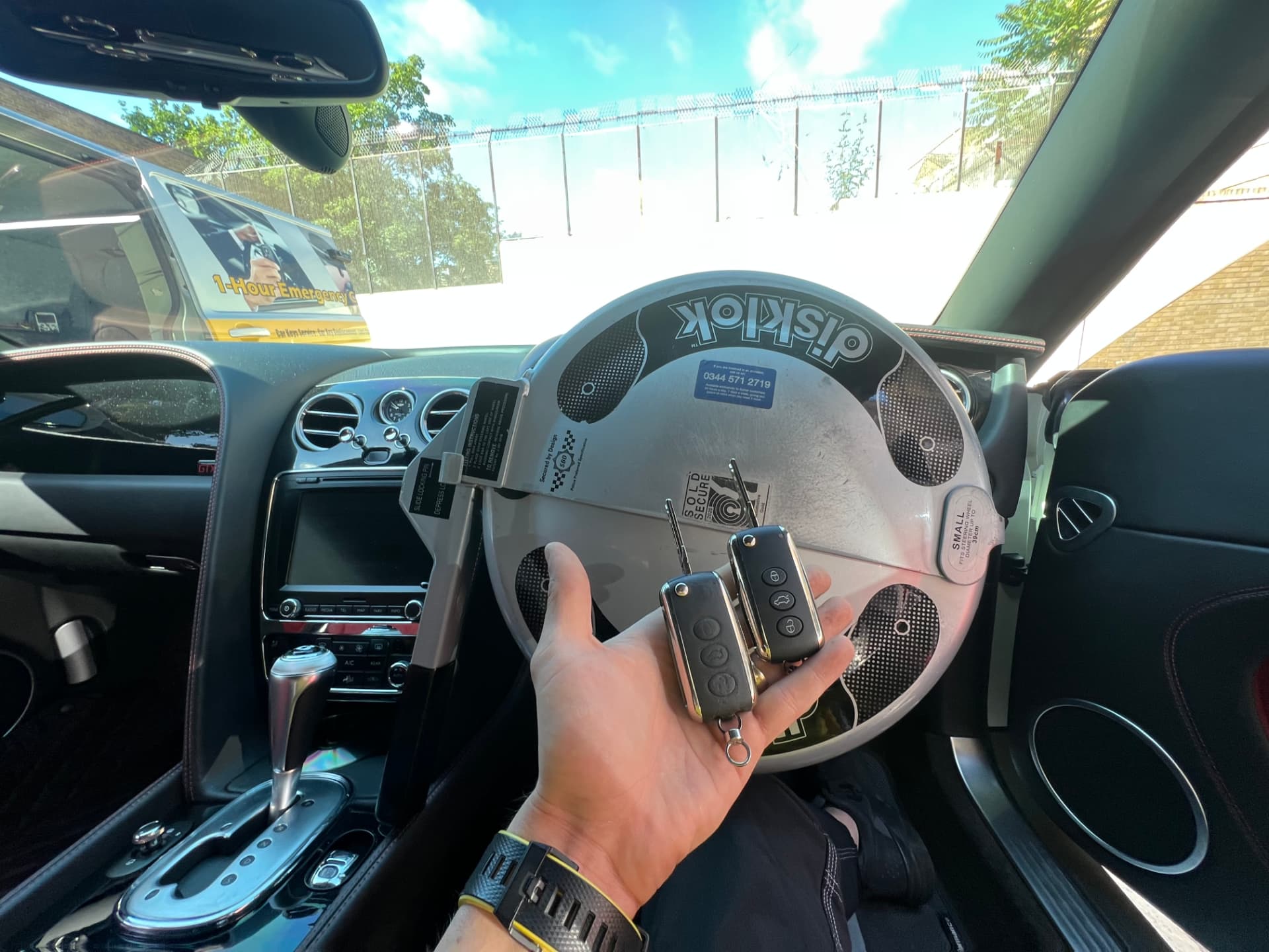Two Bentley keys held inside a Bentley cockpit with a steering wheel lock fitted