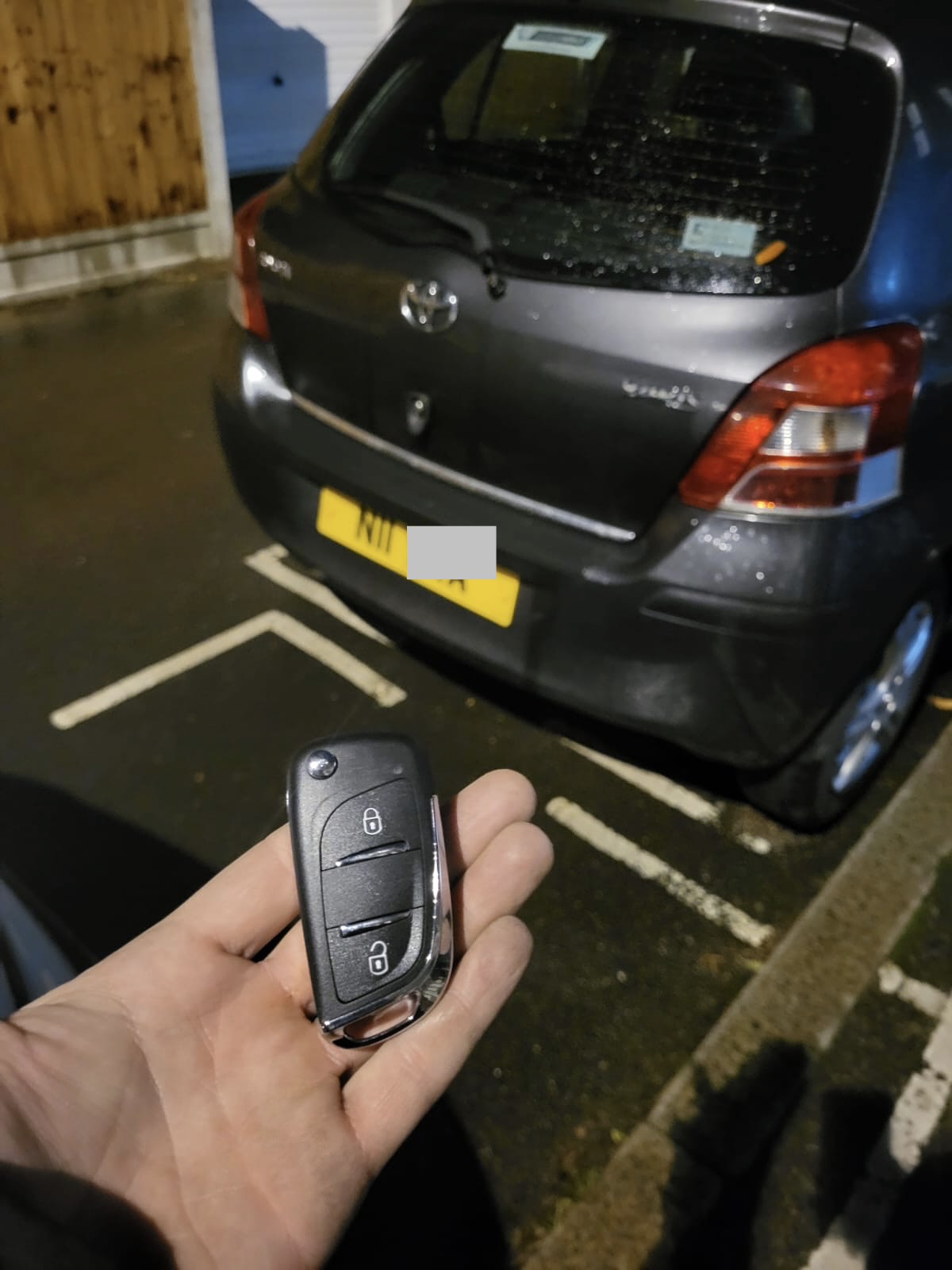 Two-button flip key held in front of a grey Toyota Yaris in a wet parking bay at night