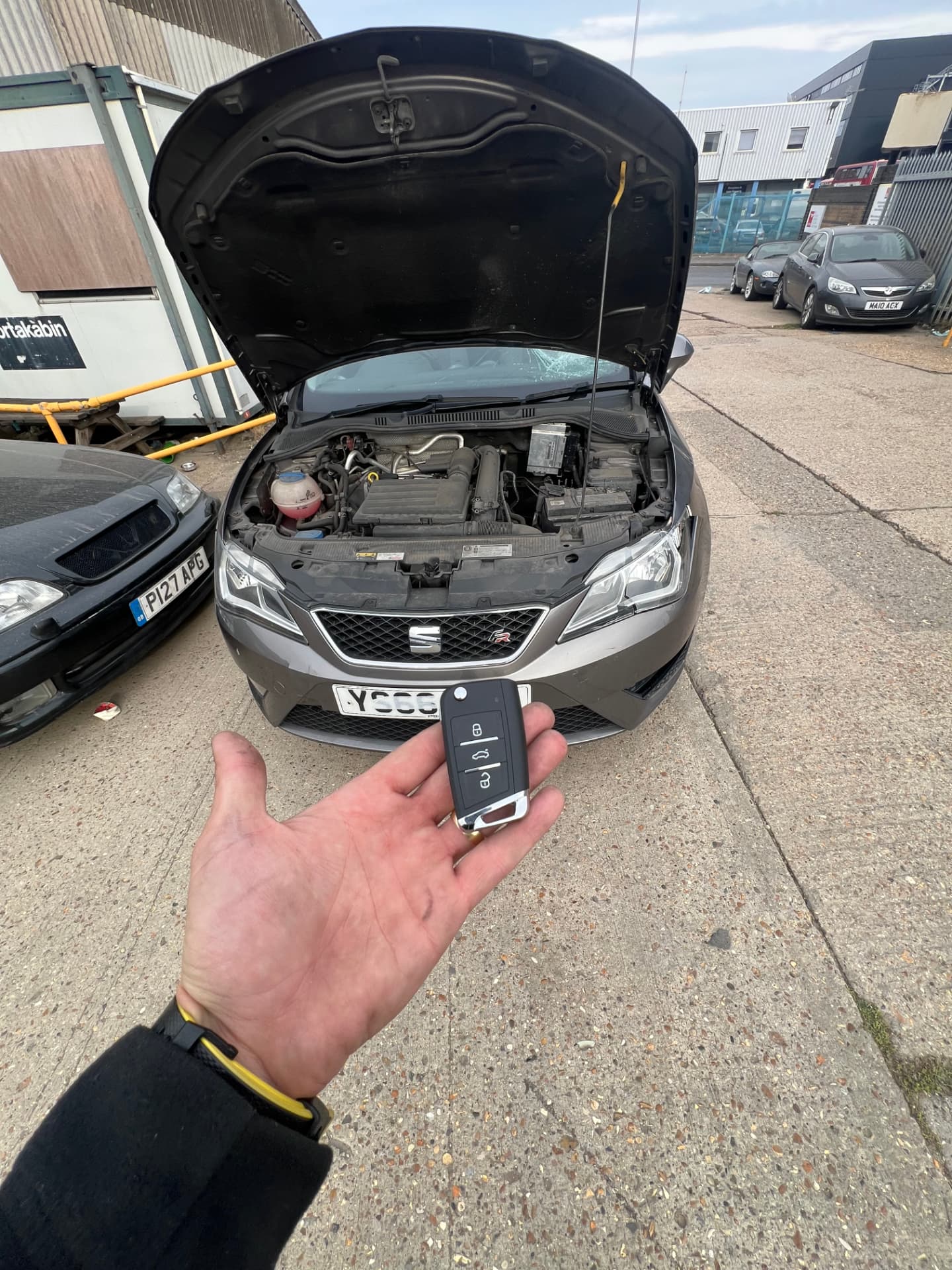 Seat flip key held in front of a grey Seat Leon FR with the bonnet open