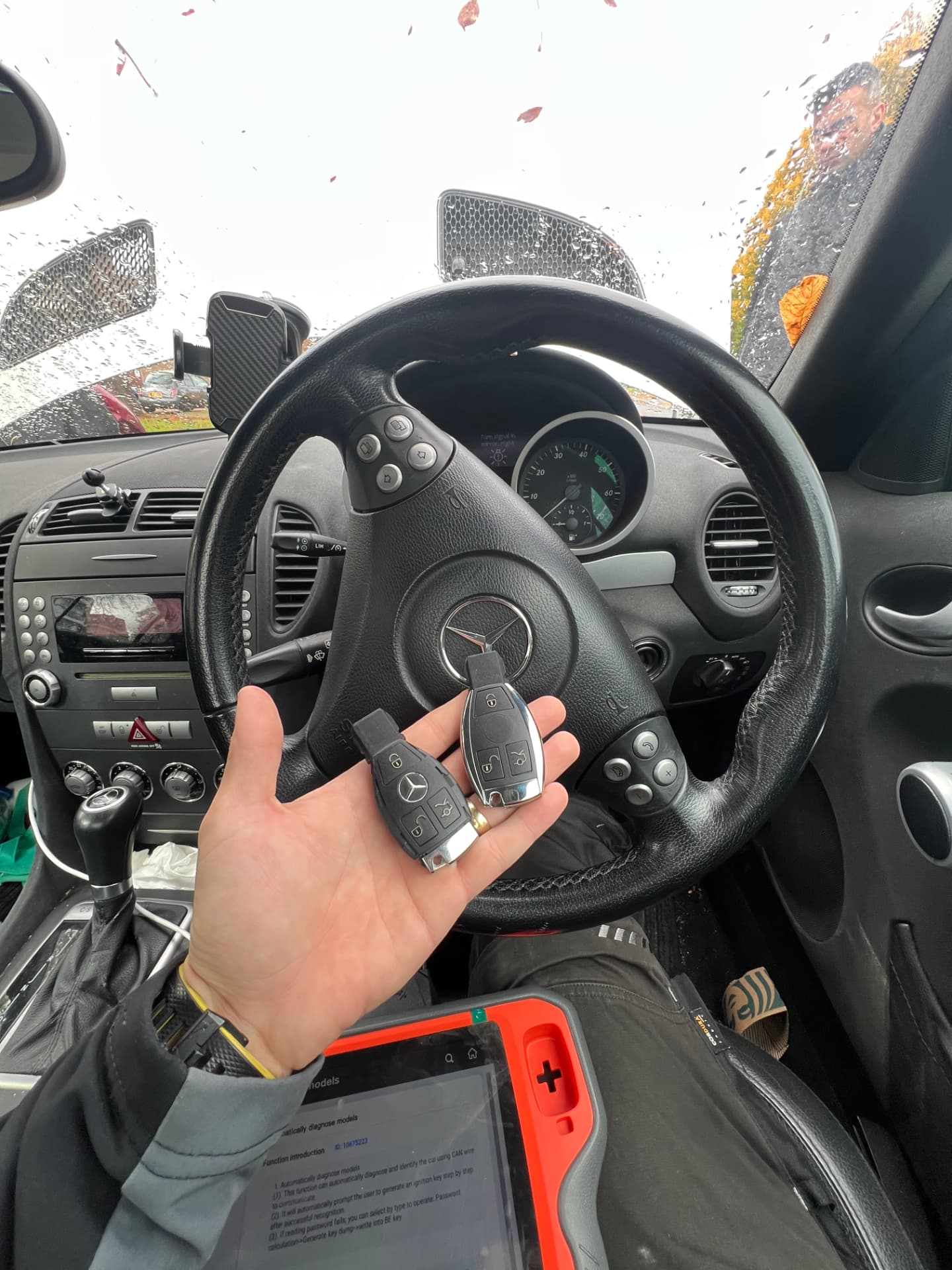 Two Mercedes keys held at the wheel of a Mercedes SLK with a diagnostic tablet on the technician's lap