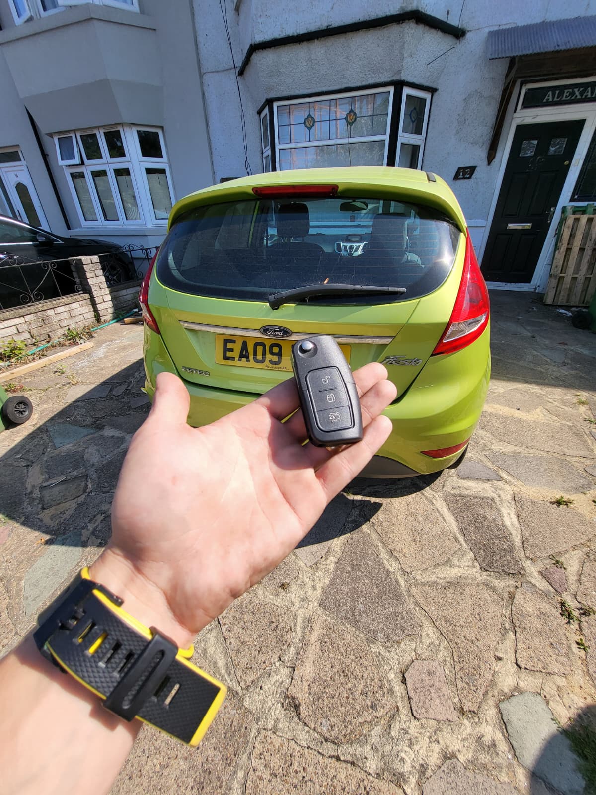 Auto locksmith holding a three-button Ford flip remote key in front of a green Ford Fiesta Zetec on a residential driveway