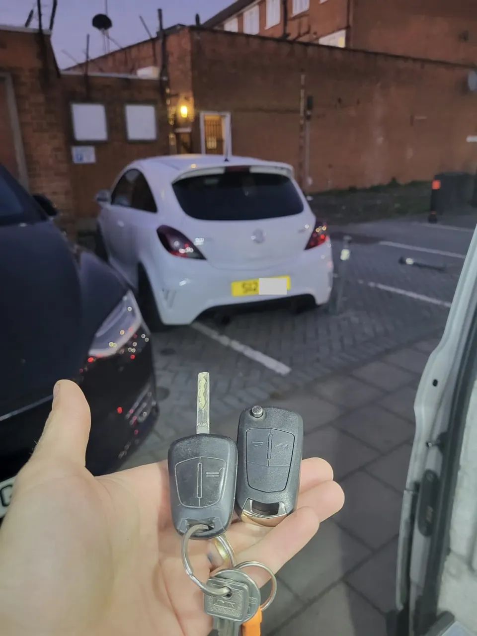 Vauxhall blade key and flip remote key held beside the locksmith van in front of a white Vauxhall Corsa