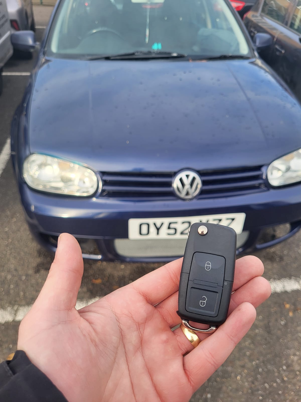 Two-button Volkswagen flip remote key held in front of a blue Volkswagen Golf in a car park