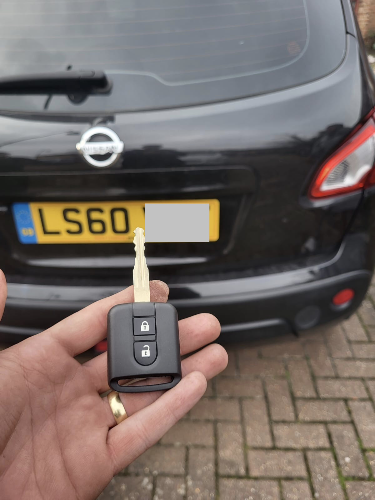 Nissan two-button remote key held in front of a black Nissan Qashqai on a block-paved drive