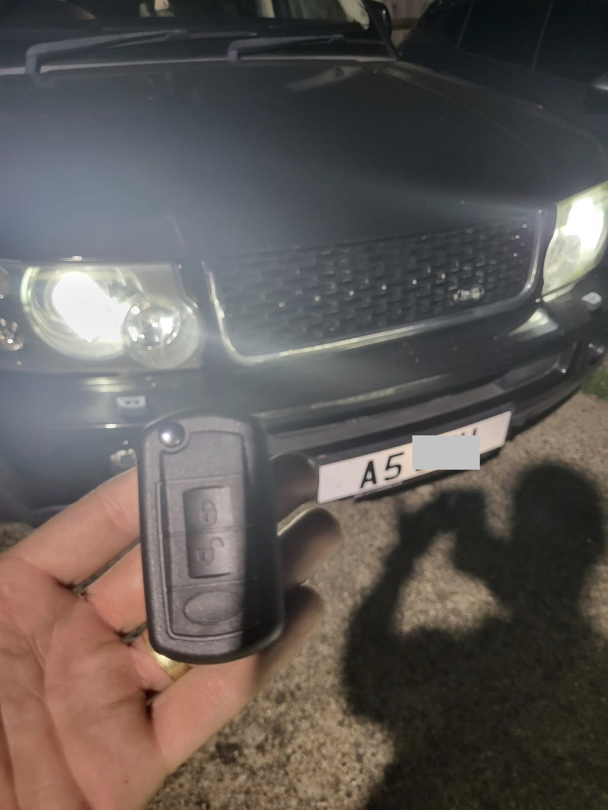 Land Rover flip remote key held in front of a Range Rover Sport with headlights on at night
