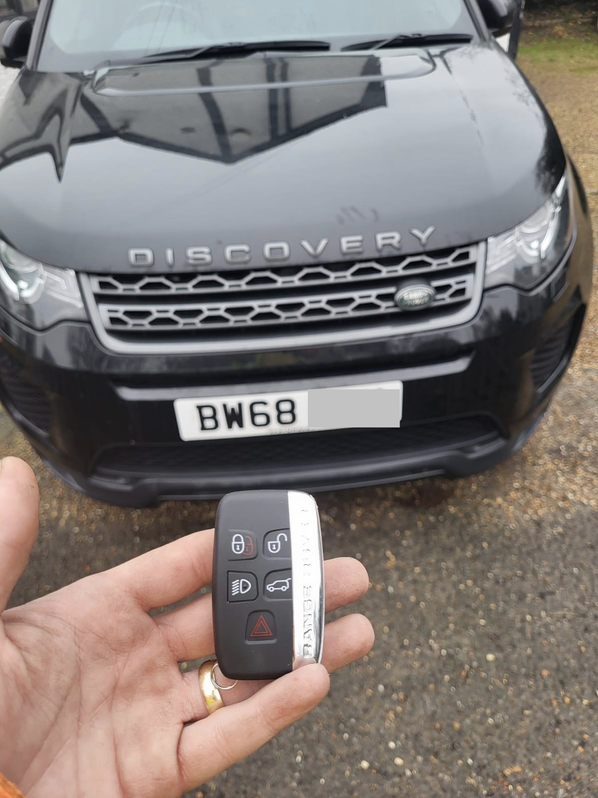 Five-button Land Rover smart key held in front of a black Land Rover Discovery Sport on a gravel drive