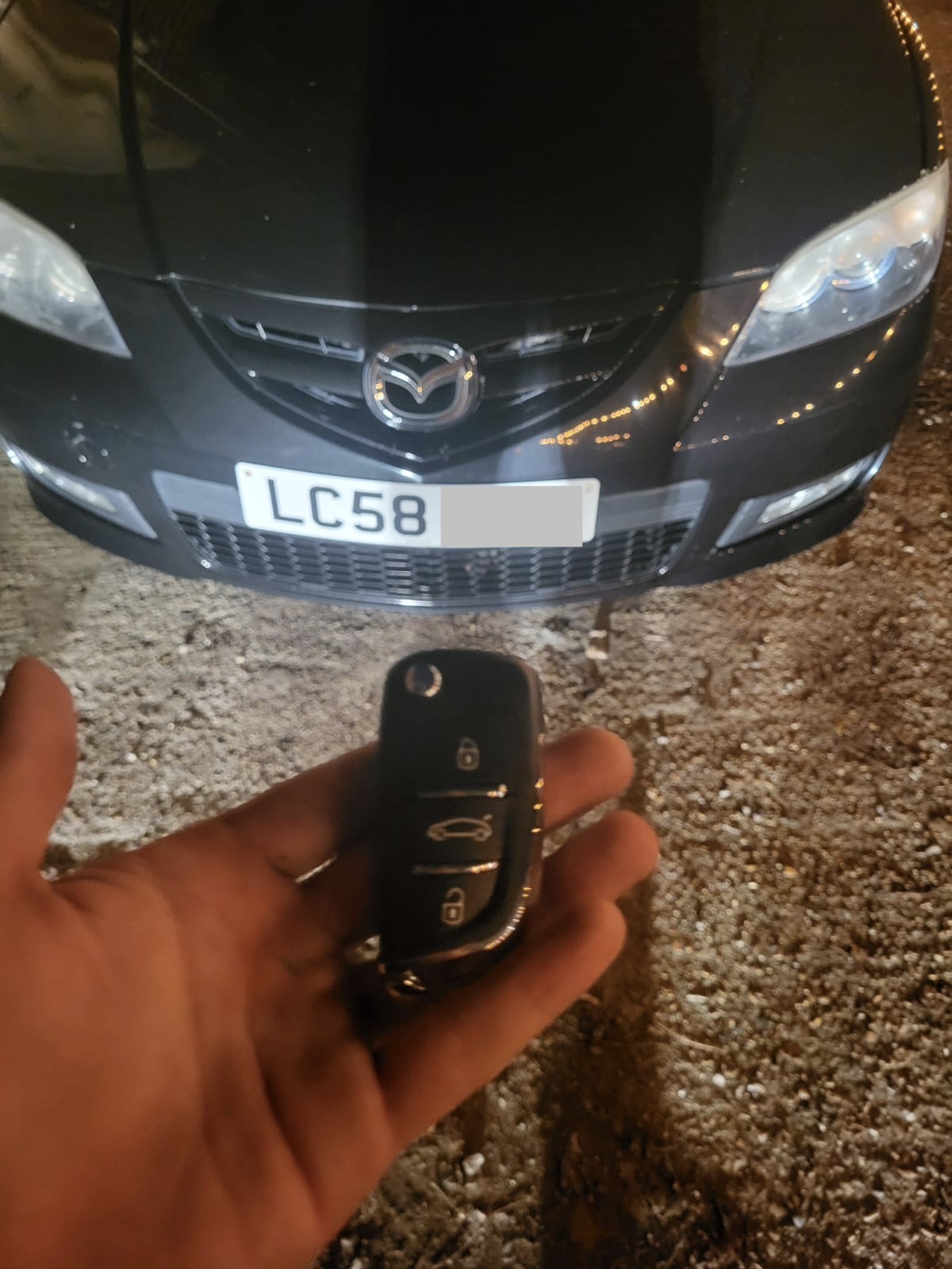 Three-button Mazda flip remote key held in front of a dark grey Mazda 3 at night