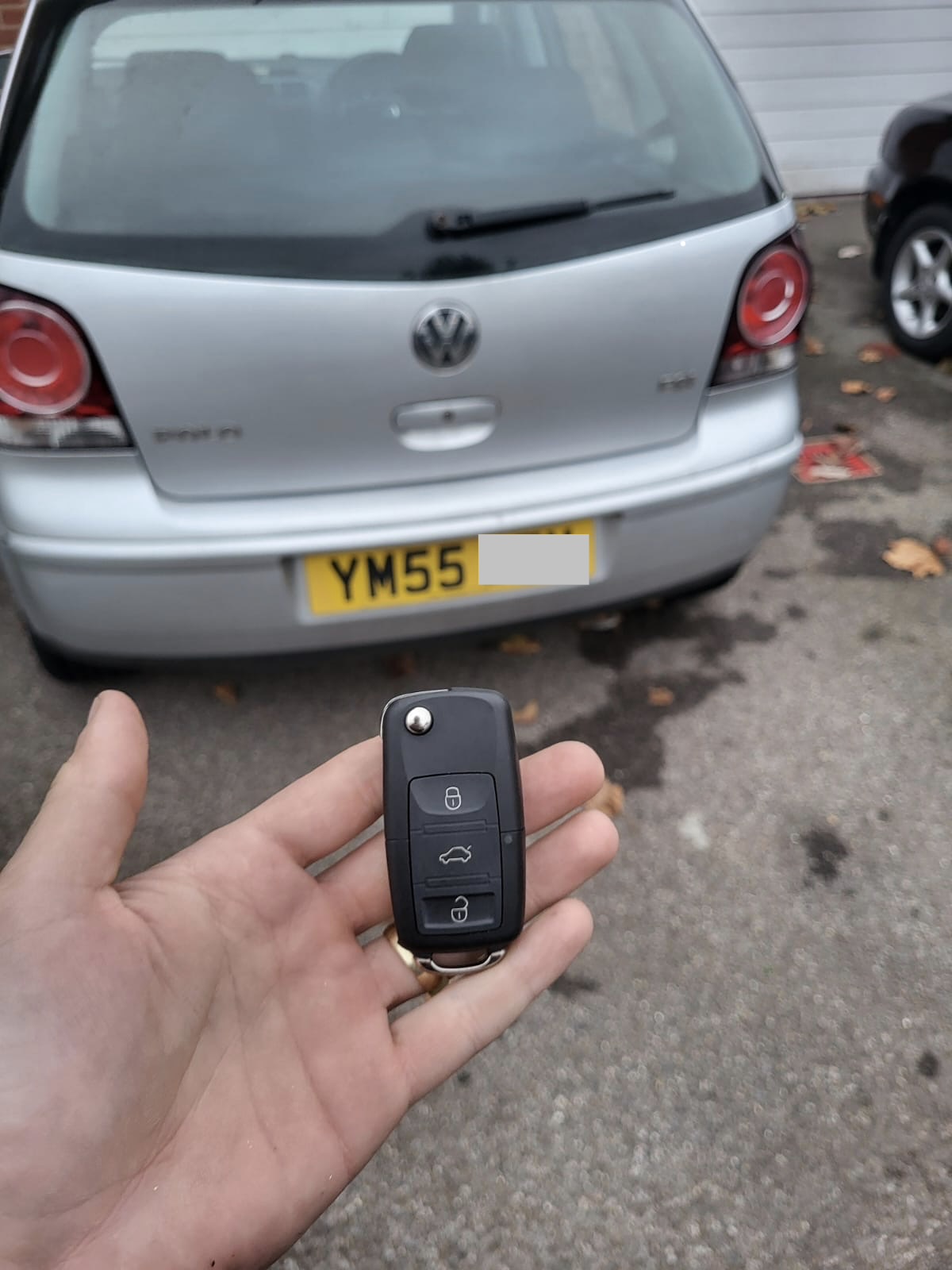 Three-button Volkswagen flip remote key held in front of a silver Volkswagen Polo
