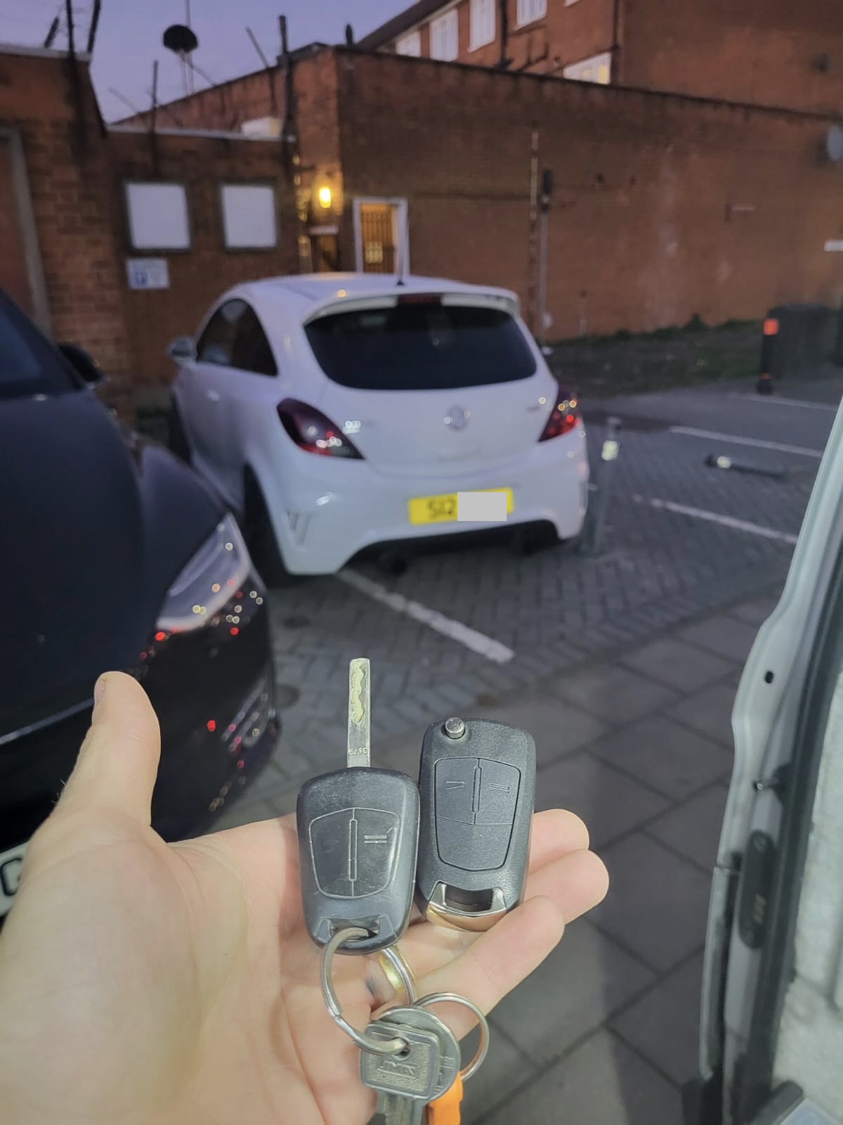Vauxhall blade key and flip remote key held beside the locksmith van in front of a white Vauxhall Corsa