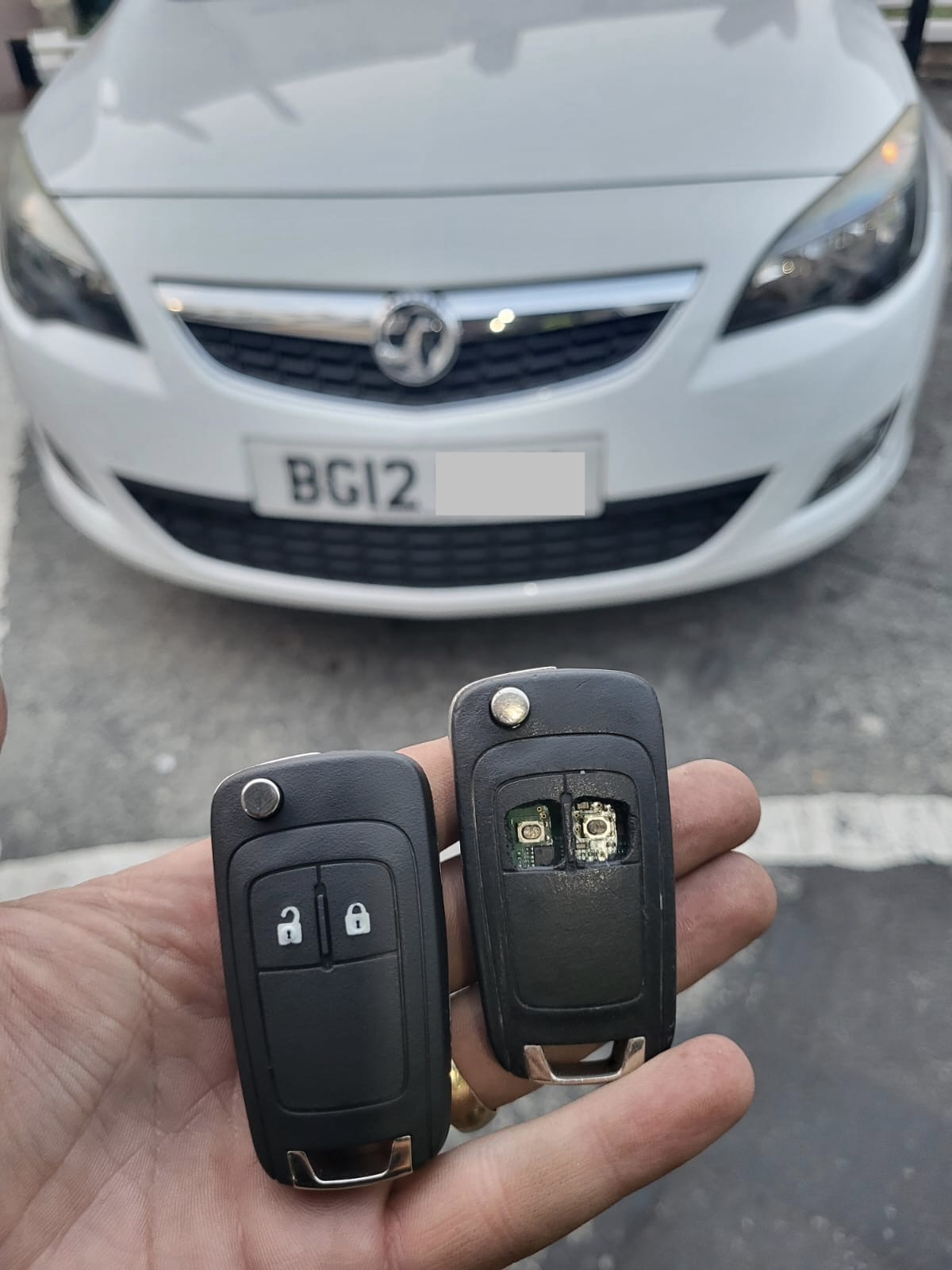 Two Vauxhall flip remote keys, one opened to show the circuit board, in front of a white Vauxhall Astra