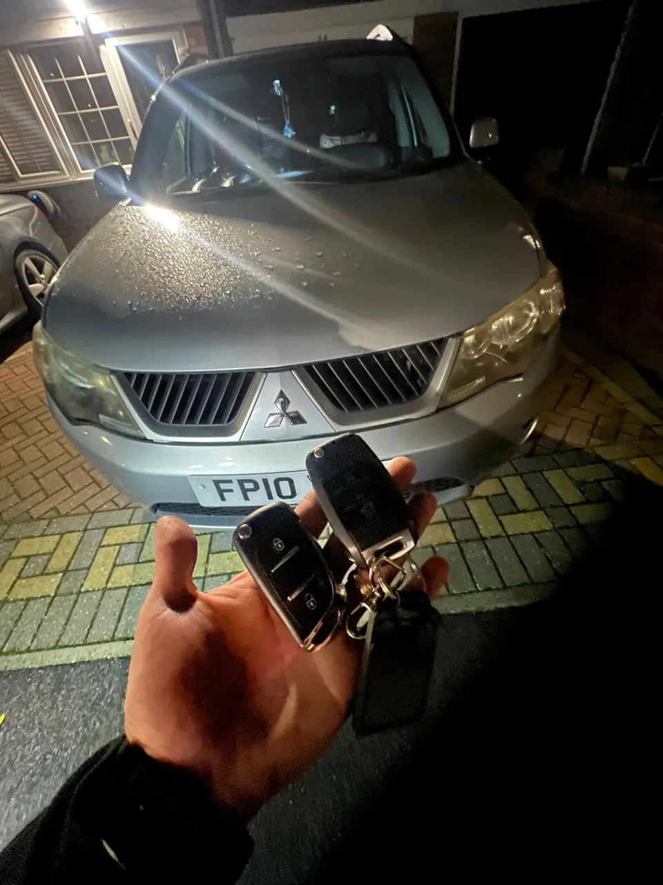 Two remote keys held in front of a silver Mitsubishi Outlander on a driveway at night